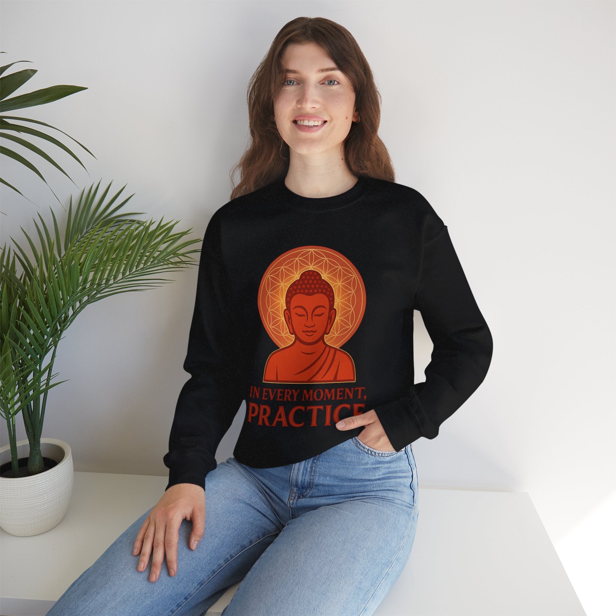 Person wearing a black sweatshirt with a Buddha graphic and text, sitting in a room with a plant.